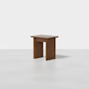 A small, modern wooden side table with a rectangular top and wide, flat legs, placed on a plain, light-colored surface against a white background.