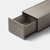 A minimalist, rectangular metallic drawer is partially open, revealing an empty interior. The drawer and its casing are finished in a matte gray color, set against a plain light background.