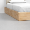A close-up view of the lower corner of a wooden bed frame with a white mattress, white sheets, and pillow, set on a light gray concrete floor.