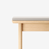 A close-up view of a light wooden bench with a soft, light gray cushion on top, set against a plain white background.