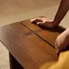 A persons hands rest on a smooth, brown wooden table with a rectangular slot in the center. The background is softly blurred.