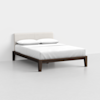 A minimalist bed with a dark wood frame, white upholstered headboard, and neatly made white bedding with two pillows, set in a plain, light-colored room.