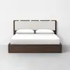 A modern wooden platform bed with a white upholstered headboard, two white pillows, and white bedding, standing against a plain white wall on a light gray floor.