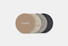 Four round felt coasters in shades of brown, beige, light gray, and dark gray are lined up. The front coaster is tan and has THUMA written in white letters. The background is plain and white.