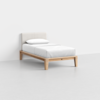 A single bed with a light wooden frame, white mattress, white pillow, and a light upholstered headboard, placed in a minimalistic, empty white room.