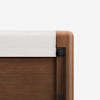 Close-up view of the corner of a wooden bed frame with a white fabric cover draped over the top edge. The wood has a smooth, polished finish and rounded edges.