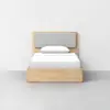 A modern wooden bed frame with a light gray upholstered headboard and white bedding, positioned against a plain white wall on a gray floor.