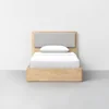 A modern wooden bed frame with a light gray upholstered headboard and white bedding, positioned against a plain white wall on a gray floor.