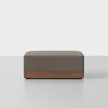 A rectangular ottoman with brown fabric upholstery and a wooden base, positioned on a light-colored floor against a plain white background.