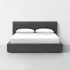 A modern dark gray upholstered bed with a low headboard, neatly made with white bedding and two white pillows, set against a plain white wall and light gray floor.