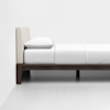 A minimalist bed with a light fabric headboard, white bedding, white pillow, and a dark wooden frame, set against a plain white wall and floor.