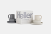 A Heller Max breakfast set is displayed with a white box labeled Heller in large letters. Two sets of stacked plates, saucers, and mugs in gray and beige are arranged beside the box on a white background.
