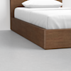 A corner view of a bed with a wooden frame and white bedding on a light gray concrete floor.