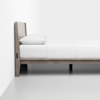 A minimalist wooden bed frame with a light finish stands against a white wall. The bed is neatly made with white bedding and a single pillow, and the room has a clean, modern aesthetic with a smooth gray floor.