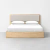 A minimalist wooden bed with a rectangular frame and a black cushioned headboard. The bed is neatly made with white bedding and two pillows, set against a plain white wall on a gray floor.