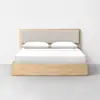 A minimalist wooden bed with a light beige upholstered headboard, two white pillows, and white bedding, placed against a plain white wall on a gray floor.