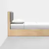 A minimalist wooden bed with a tall headboard, featuring two gray cushions, white bedding, and a pillow, placed against a plain white wall and gray floor.