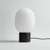 A modern table lamp with a matte black cylindrical base and a smooth, rounded white glass shade, placed on a white surface with a black power cord extending to the right.