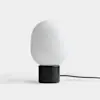 A modern table lamp with a matte black cylindrical base and a smooth, rounded white glass shade, placed on a white surface with a black power cord extending to the right.