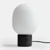 A modern table lamp with a matte black cylindrical base and a smooth, rounded white glass shade, placed on a white surface with a black power cord extending to the right.