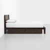 A dark wooden bed frame with a headboard, white bedding, and two under-bed storage drawers, positioned against a white wall in a minimalist room with a light gray floor.