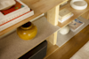A close-up of a wooden bookshelf displaying art books, a yellow vase, a white round sculpture, a black speaker, and a few smaller books, with a textured beige shelf and wood flooring visible.