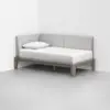 A modern daybed with a light gray upholstered backrest and a wooden frame, placed in the corner of a minimalist room with white walls and a light gray floor. A white pillow and bedding are neatly arranged on the bed.