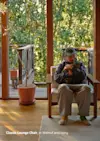 A person wearing glasses and a cap sits in a wooden lounge chair, holding a cat. Sunlight streams through large windows, revealing greenery and a deck outside. A potted plant is beside the chair. Text reads: Classic Lounge Chair, in Walnut and Ivory.