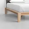 Close-up of a wooden bed frame with light-colored wood, showing the corner and legs on a smooth, light gray floor. White bedding and a pillow are neatly arranged on the bed.