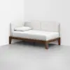 A modern daybed with a wooden frame, white mattress, and white upholstered back and side rests, positioned in the corner of a minimalist room with light gray flooring and white walls. A single white pillow rests on the bed.