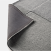 A close-up view of a gray textured rug with a corner folded over, showing its dark backing and stitched edge details. The rug is placed on a light background.