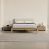 A modern minimalist bedroom features a wooden bed with a light gray upholstered headboard, white bedding, two matching wooden nightstands, a small tree branch decor, a lamp, and a rug on a light floor.