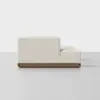 Side view of a modern, light beige upholstered sofa with a low back and seat, featuring a wooden base, placed on a plain light gray floor against a white background.