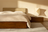 A minimalist bedroom with a wooden bed frame, beige bedding, and a matching headboard. A wooden nightstand holds a sculptural lamp and a cup. Soft sunlight streams in, casting shadows on the light wall and rug.