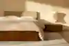 A minimalist bedroom with a wooden bed frame, beige bedding, and a matching headboard. A wooden nightstand holds a sculptural lamp and a cup. Soft sunlight streams in, casting shadows on the light wall and rug.