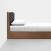 A minimalist wooden bed frame with a black cushioned headboard, white pillow, and white bedding, set against a plain white wall and gray floor.