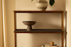 A wooden shelf against a beige wall holds a ceramic vase with green leaves on the top, a dark bowl on the middle shelf, and a stack of books with a small cup on the bottom shelf.