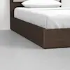 A close-up of the corner of a modern bed with a dark wooden frame, white bedding, and a white pillow, set on a light gray floor.