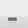A minimalist wooden coffee table with a single open drawer sits on a smooth, light gray floor against a plain white background.