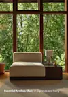 A modern armless chair in ivory upholstery and espresso base sits beside a dark side table with a lamp and book, placed in front of large windows overlooking lush green trees. Text reads: Essential Armless Chair, in Espresso and Ivory.