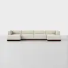A modern, modular beige sectional sofa with low backs and arms, set on a dark wooden base, displayed in a minimal, light gray room with a plain white background.