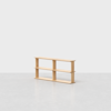 A minimalist, light wood bookshelf with three shelves and an open-back design, placed in a bright, empty room with a smooth, pale gray floor and a plain white background.