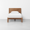 A single wooden bed with a simple headboard, white bedding, and a white pillow, positioned against a plain white wall on a light gray floor.