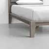 Close-up of a wooden bed frame with a neatly made bed featuring white sheets and pillow, set on a light gray floor against a white wall.