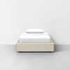 A single beige upholstered bed with white sheets and a single pillow, placed against a plain white wall on a light gray floor, viewed from the front.