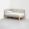 A minimalist daybed with a beige upholstered backrest, a wooden frame, and a single white pillow and sheet, positioned in the corner of a plain white room.