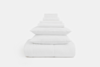 A neatly stacked set of white bedding essentials, including a comforter, pillows, and folded sheets and towels, arranged in descending size order against a plain white background.