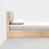 A modern wooden bed with a light wood frame, beige padded headboard, white pillow, and white bedding, set against a plain white wall and gray floor.