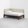 A minimalist daybed with beige upholstered back and side panels, a dark wooden frame, a white mattress, and a single white pillow, positioned in the corner of a plain, light-colored room.