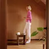 A woman with blonde hair, wearing a pink sweater and white pants, stands barefoot beside a small wooden table with a candle and a book in a warmly lit, minimalist room with wood-paneled walls and a large green plant.